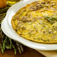 Frittata di asparagi selvatici dorata su piatto bianco, con asparagi freschi e uova sullo sfondo.