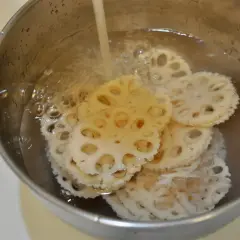Slices of pickled lotus root are being rinsed in a metal bowl under running water, ready for a delicious meal.