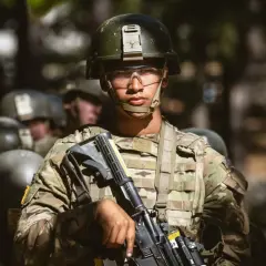 A young soldier, determined and focused, stands ready during far bootcamp. He wears a helmet, protective eyewear, and camouflage fatigues, holding his rifle.
