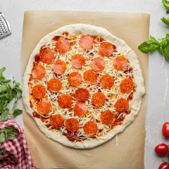 Pizza z sosem, serem i okrągłymi plasterkami pepperoni, gotowa do pieczenia. Obok świeża bazylia i pomidorki.