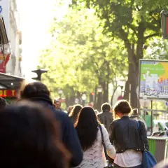 Des passants se promènent sur un grand boulevard parisien, sous un soleil radieux. Un panneau "METRO" rouge indique une station.
