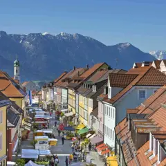 Bunte Häuserfronten und ein Markt beleben die Gasse in Murnau am Staffelsee. Im Hintergrund ragen schneebedeckte Berge auf.