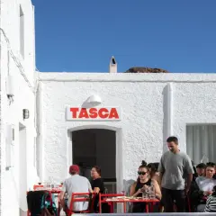 Gente disfrutando en el bar Tasca, con sus altas hortichuelas, bajo un cielo azul.