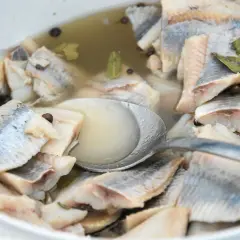Kawałki śledzi w zalewie z przyprawami, gotowe do podania. Widać łyżkę zanurzoną w marynacie.