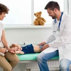 Lekarz bada brzuszek dziecka, które leży na kozetce. Mama z pluszowym misiem obok. Dziecko uśmiecha się, mimo że może mieć biegunkę u niemowlaka.