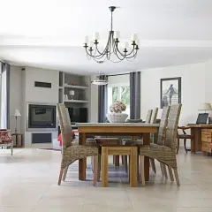 Elegancki, czarny żyrandol do salonu nad stołem z wiklinowymi krzesłami. W tle kominek i wygodne fotele.