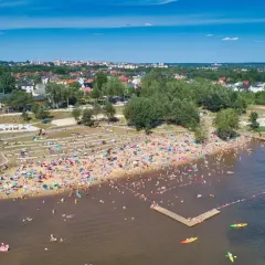 Tłum ludzi wypoczywa na plaży i kąpie się w **Zalewie Lubianka**. Widać molo, dmuchane atrakcje wodne i kajaki.