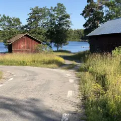 Väg leder mot Långvinds bruk med röda byggnader vid en sjö. Sommarängar och träd.