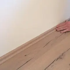 Hand drückt Sockelleisten an die Wand. Der Boden ist aus hellem Holz.