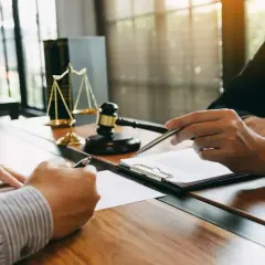 Dwóch mężczyzn przy biurku, jeden podpisuje dokument, drugi wskazuje piórem. W tle waga sprawiedliwości i młotek. Po co notariusz przy wynajmie mieszkania? By mieć pewność prawną.