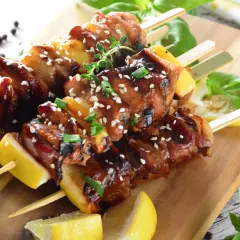 Aromatyczne yakitori z kurczaka w sosie teriyaki, posypane sezamem i szczypiorkiem, podane z cytryną na drewnianej desce.