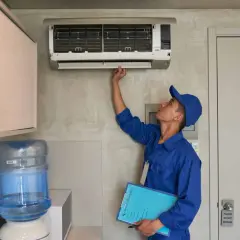 Un technicien en uniforme bleu vérifie une installation de climatisation murale, tenant un dossier de service.