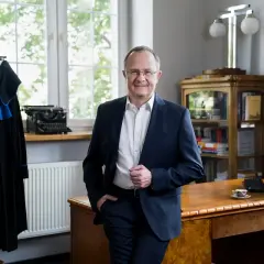 Mężczyzna w garniturze stoi w gabinecie, obok wieszaka z togą prawniczą.
