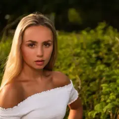 Piękna fotografia portretowa młodej kobiety z długimi blond włosami, w białej bluzce, na tle zielonej natury.