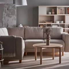 Przytulny salon szaro beżowy inspiracje: wygodna kanapa, okrągły stolik, regał z dekoracjami i kosz z tkanin.