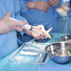 Dłonie w rękawiczkach chirurgicznych trzymają strzykawkę nad tacą z narzędziami i miskami.
