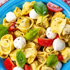 Ein bunter Tortellini Salat mit Mozzarella-Kugeln, Tomatenstücken und frischem Basilikum, appetitlich in einer blauen Schüssel angerichtet.