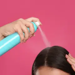 Dłoń trzymająca turkusowy spray z suchym szamponem, rozpylająca go na ciemne włosy.