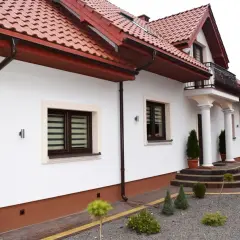 Biały dom z brązowymi rynnami i dachówką. Eleganckie cokoły podkreślają jego architekturę.