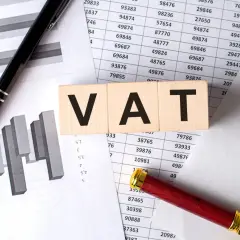 Drewniane klocki z napisem VAT leżą na dokumentach z wykresami i tabelami. Jak odliczyć VAT z faktury? Pióro i lupa sugerują analizę.