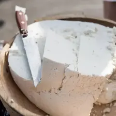Formaggio fresco e morbido, simile a uno stracchino, tagliato in una ciotola di legno con un coltello.