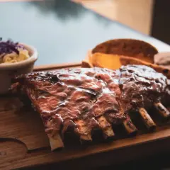 Délicieuses côtes levées au bbq, nappées de sauce, servies avec une salade de chou et des quartiers de pommes de terre sur une planche en bois.