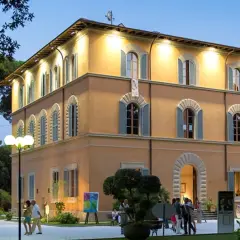 Edificio giallo illuminato nel parco della Versiliana, con persone che passeggiano e un'opera d'arte esposta.