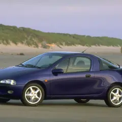 Una donna appoggiata a una Opel Tigra blu, icona delle macchine anni 90, sulla spiaggia al tramonto.