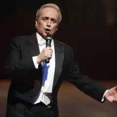 José Carreras, hiszpański tenor, w eleganckim smokingu śpiewa do mikrofonu, gestykulując.