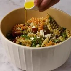 Un bol lleno de pasta de colores, tomates cherry, queso y rúcula, con aceite de oliva vertiéndose sobre ella.
