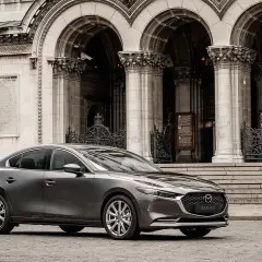 Elegancka, szara Mazda 3 2025 przed monumentalną, kamienną budowlą z arkadami i ozdobnymi bramami.