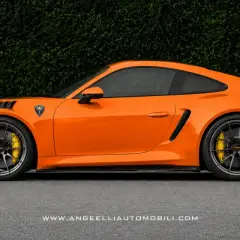 Pomarańczowy Porsche RWB z czarnym spoilerem i felgami, na tle zielonego żywopłotu.