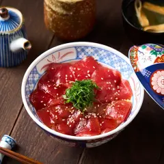 A bowl of delicious Japanese sweet rice topped with thinly sliced tuna, sesame seeds, and green garnish. A decorative lid rests beside it.