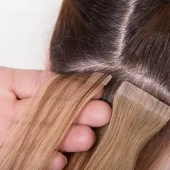 Manos aplicando extensiones adhesivas rubias a cabello castaño. El proceso de colocación de extensiones adhesivas es delicado.