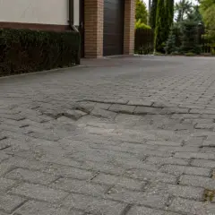 Uszkodzona kostka brukowa przy drzwiach wejściowych do garażu. Widać zapadnięte i przesunięte fragmenty nawierzchni.