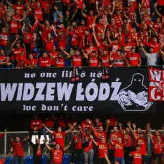 Tłum kibiców Widzewa w czerwonych koszulkach skanduje przyspiewki na Widzew, machając rękami. Na banerze widnieje napis "WIDZEW ŁÓDŹ".