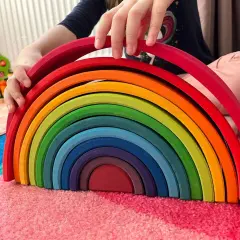 Dziecko bawi się tęczowym łukiem, jedną z popularnych zabawek, układając kolorowe drewniane elementy na dywanie.