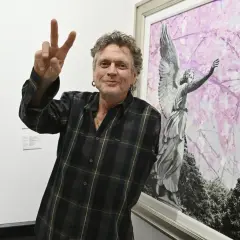 Rick Allen, perkusista Def Leppard, pozuje z uniesionym znakiem pokoju obok obrazu z aniołem i kwitnącymi drzewami.