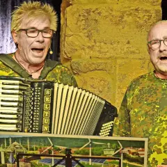 Zwei Männer im Tarnanzug singen und spielen Akkordeon beim **Kabarett der Galgenstricke**. Ein Bild mit Stadtansicht und Windrädern liegt vorn.