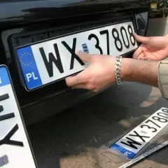 Montaż nowych tablic rejestracyjnych. Jedna z nich, z numerem WX 37808, jest właśnie wkładana. Obok leży kolejna, zapakowana.