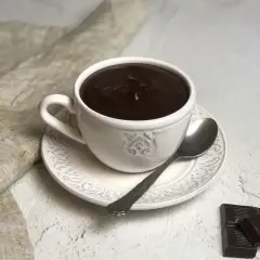 Una tazza di cioccolata calda cremosa, perfetta per imparare come fare una buona cioccolata calda. Accanto, due quadratini di cioccolato fondente e un cucchiaino.