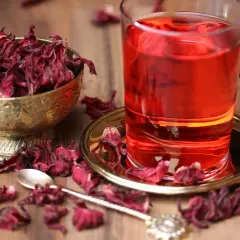 Ein Glas leuchtend roter Malva Sylvestris Tee steht neben einer Schale getrockneter Blüten. Getrocknete Blütenblätter sind auf dem Holztisch verstreut.
