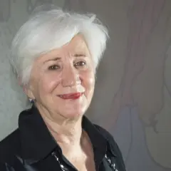 Olympia Dukakis ler vänligt mot kameran, med sitt karakteristiska vita hår och en svart skjorta. Bakom henne anas en tecknad figur.