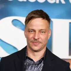 Tom Wlaschiha, mit einem Schnurrbart und blauen Augen, posiert vor einem Hintergrund mit großen weißen Buchstaben. Er denkt vielleicht an seine tom wlaschiha kinder.