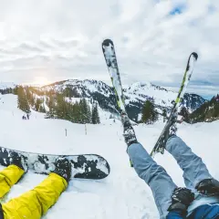 Dwie osoby odpoczywają na śniegu. Jedna leży na desce snowboardowej, druga ma założone narty. W tle góry i stoki narciarskie.