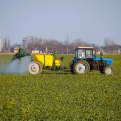Niebieski traktor z żółtym zbiornikiem wykonuje oprysk na polu zielonych roślin. W tle widać przemysłowe budynki.