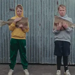 Två barn, som påminner om Sverrir Gudnason som barn, håller varsin fisk framför en plåtvägg.