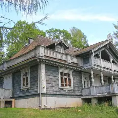 Drewniany dworek w parku pałacowym w Białowieży, z werandami i ozdobnymi detalami, otoczony zielenią.