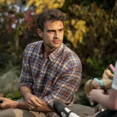 Theo James w kratowanej koszuli, znany z filmów, seriali i programów, siedzi na zewnątrz. Obok niego dziecko bawi się lalką.