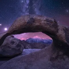 A natural rock arch frames a majestic mountain range under a breathtaking starry sky.